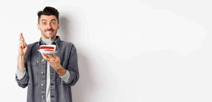 Birthday. Happy guy making wish with fingers crossed, holding bday cake with candle, standing on white background.