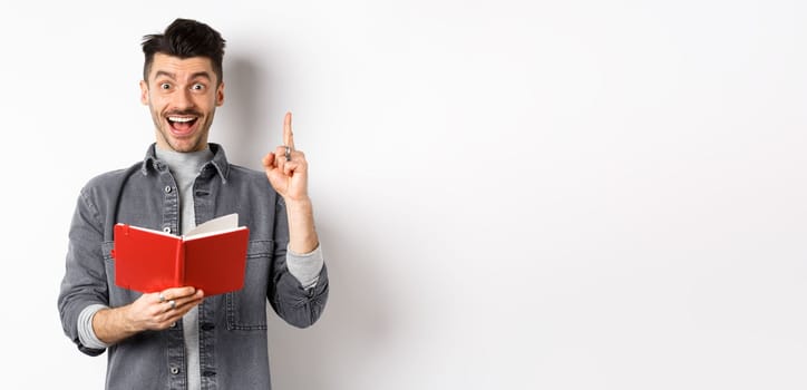 Excited guy pitching an idea while doing homework, raising finger and holding planner, standing against white background.