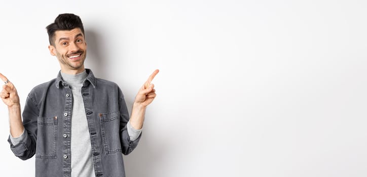 Young handsome guy with moustache pointing fingers sideways, showing two logos, smiling happy at camera, standing in casual clothes on white background.