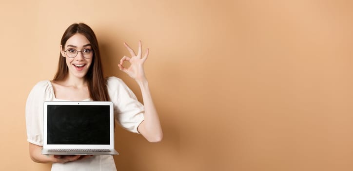 E-commerce. Excited young woman in glasses showing okay sign and laptop screen, recommending internet promo, standing on beige background.