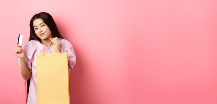 Shopping. Stylish asian girl showing shop bag and plastic credit card, standing on pink background.