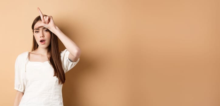 Confident woman mocking people with loser sign, being mean, standing on beige background.