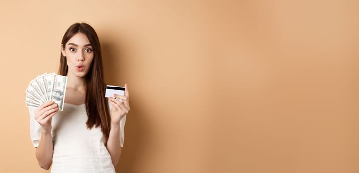 Excited girl showing dollar bills and plastic credit card, saying wow with amazed face, standing on beige background.