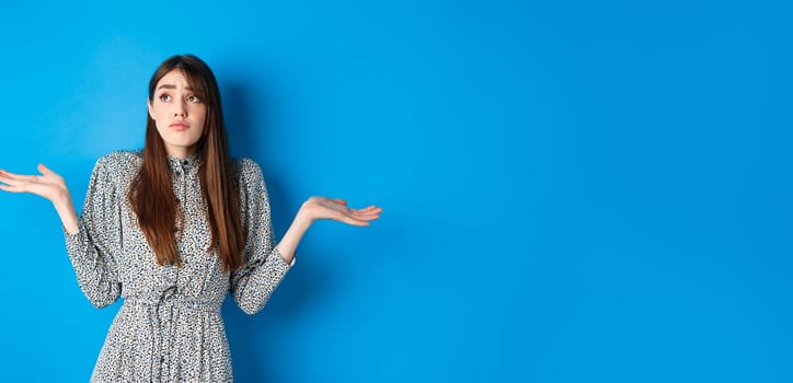 Confused pretty girl in dress shrugging shoulders and looking aside sad, know nothing, standing against blue background.