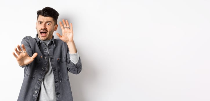 Scared timid guy screaming and raising hands in pleading pose, begging to stop, defending from assault, standing on white background.