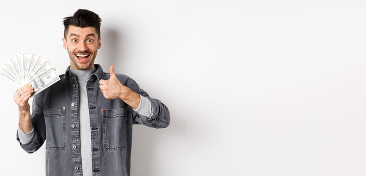 Image of excited man holding dollar bills and show thumbs-up with happy face, making money, standing on white background.