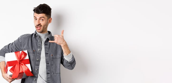 Macho guy with gift box show call me phone gesture, flirting with you, standing boastful on white background. Relationship and Valentines day concept.