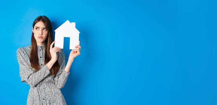 Real estate. Sad frowning girl looking aside with unfair and upset face, showing paper house cutout, standing on blue background.