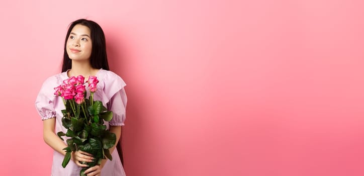Asian teenage girl in cute dress looking romantic at empty space logo, holding valentines day flowers gift, receive boquet of roses from lover, standing on pink background.