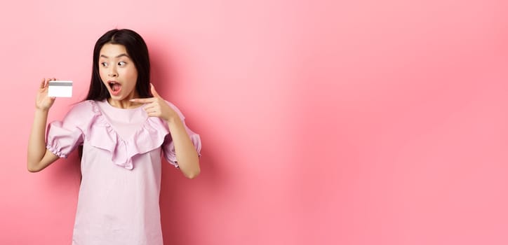 Excited asian woman pointing at plastic credit card, showing awesome advertisement, standing on pink background.