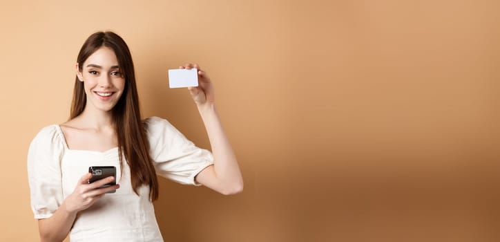 E-commerce concept. Smiling woman showing plastic credit card while shopping online on mobile phone, standing against beige background.