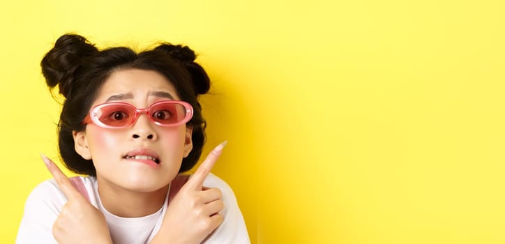 Summer fashion concept. Headshot of worried indecisive asian girl, pointing fingers sideways and showing two ways, cant choose, wearing sunglasses.