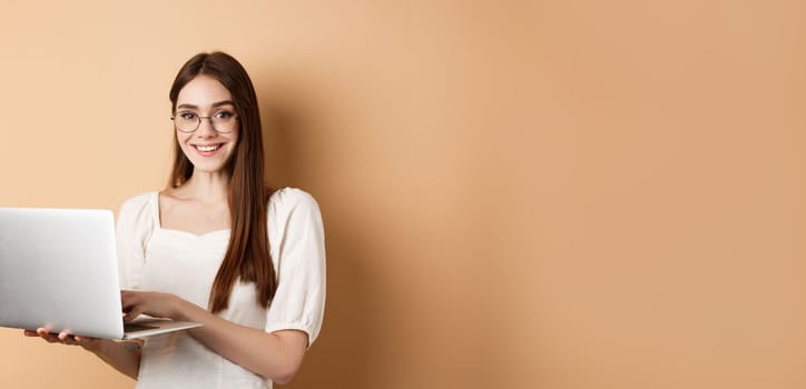 Smiling girl student working on laptop, wearing glasses and looking happy, using computer while standing against beige background.