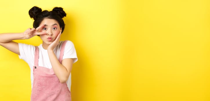 Kawaii asian girl showing v-sign and pouting cute, making silly face with makeup, standing on yellow background.