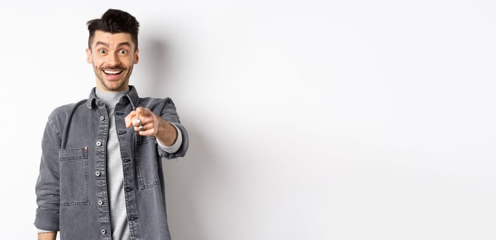 It is you. Excited smiling man pointing fingers at camera with cheerful face, recognize person, standing in denim jacket on white background.