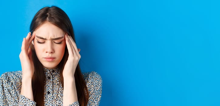 Close-up portrait of young woman feel sick, touching head temples and frowning from headache, having migraine, standing on blue background.