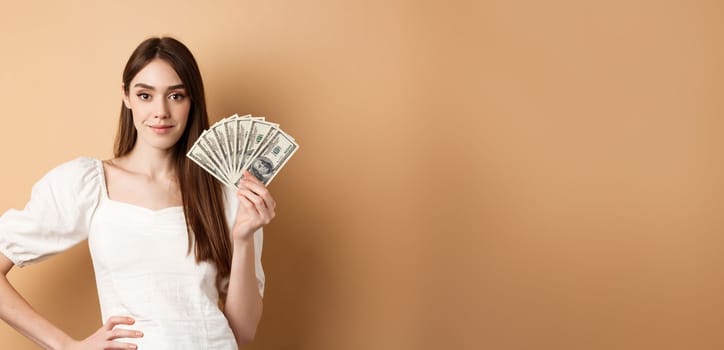 Confident smiling woman showing dollar bills, earn money and look satisfied, standing on beige background with cash.