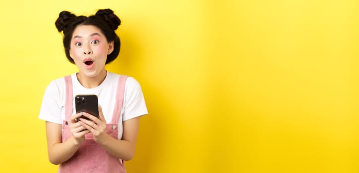 Excited girl with glam makeup and summer clothes, gasping amazed, checking out internet offer on mobile phone, standing on yellow background.