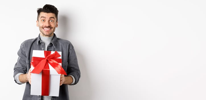 Cheerful caucasian guy holding surprise gift, receive present on holiday and smiling thankful, looking grateful at camera, celebrating Valentines day, white background.