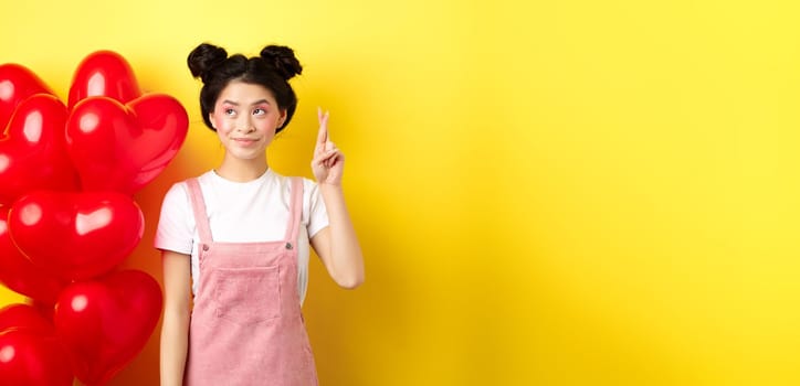 Valentines day concept. Beautiful korean girl dreaming of perfect date, making wish and looking at upper left corner, standing near red hearts balloons, yellow background.