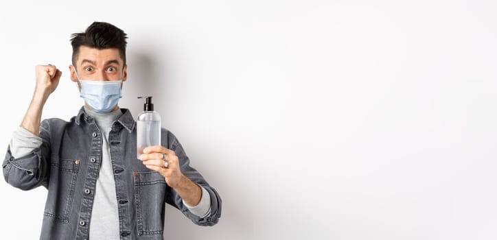 Health, covid and pandemic concept. Happy guy motivated to get rid from virus germs, showing bottle of hand sanitizer and fist pump, cheering while standing on white background.