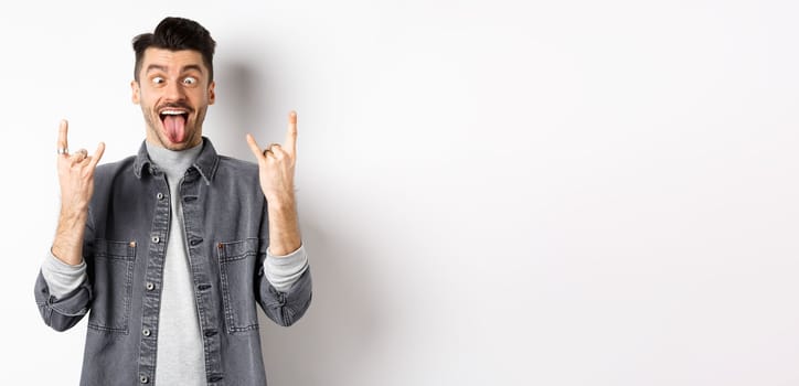 Funny young man showing tongue and rock horns, squinting eyes to fool around, enjoy party or concert, standing on white background.