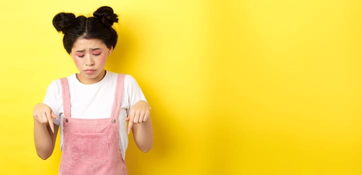 Sad miserable girl with glamour bright makeup, looking and pointing down upset, showing bad thing, standing against yellow background.