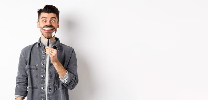 Handsome positive guy showing white perfect smile with magnifying glass, and squinting eyes, making funny faces, white background.