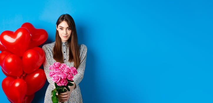 Valentines day. Pretty woman making romantic gift to girlfriend, stretch out hands with beautiful pink flowers and smiling, standing on blue background.