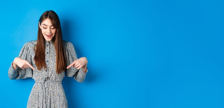 Dreamy happy woman pointing fingers down and looking wondered at promotion, showing awesome news, standing in dress on blue background.