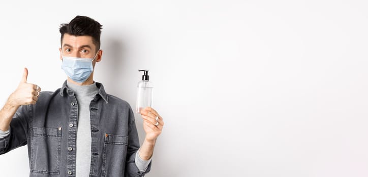 Healthy people and covid-19 concept. Excited man in sterile medical mask holding bottle of good hand sanitizer, show thumb up, recommend antiseptic, standing against white background.