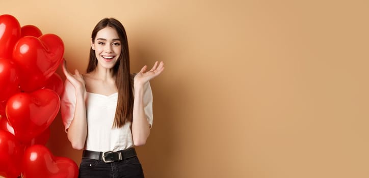 Valentines day and love concept. Happy smiling woman raising hands up surprised, standing near romantic heart balloons on beige background.