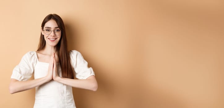 Smiling girl in glasses say thank you, show namaste gesture, express gratitude, standing pleased on beige background.