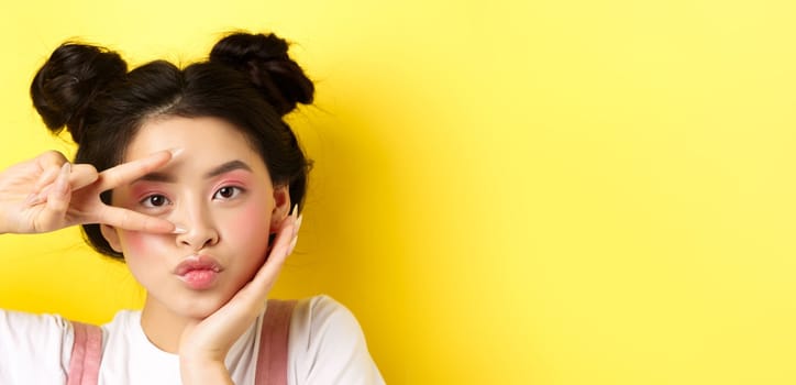 Close-up of cute asian woman with bright glamour makeup, long fingernails, showing v-sign and pouting silly at camera, standing kawaii on yellow background.