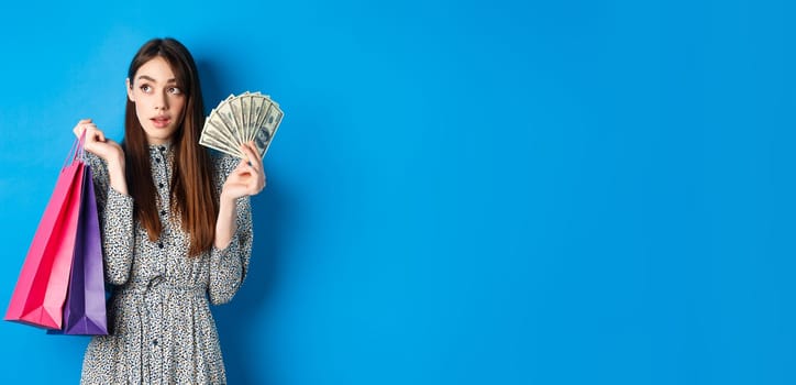 Shopping. Pretty woman thinking what to buy, looking left at logo, holding paper shop bags and money, standing on blue background.