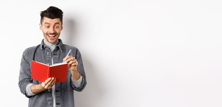 Excited guy writing in planner and smiling, writing down ideas in journal or diary, standing on white background.