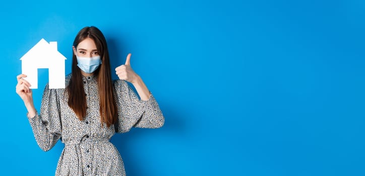Real estate, covid-19 and pandemic concept. Candid woman in medical mask showing thumb up and paper house cutout, reanting apartment, standing on blue background.