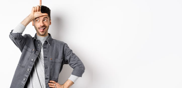 Funny guy mocking person, showing tongue and loser sign on forehead, making fun of lost team, standing on white background.