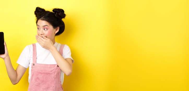 Online shopping concept. Silly japanese girl with beauty makeup, cover mouth with hand laughing and showing empty smartphone screen, show funny thing on phone, yellow background.