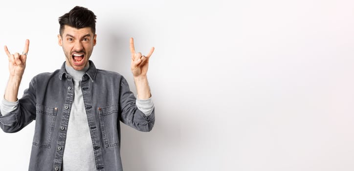 Rock on. Sassy young man shouting and showing heavy metal horns sign, attend awesome concert, having fun, standing on white background.