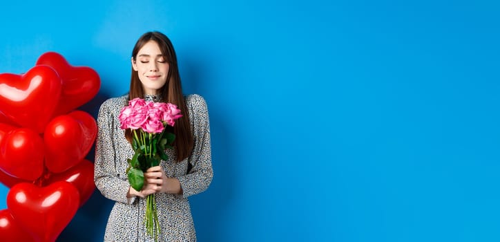Valentines day. Romantic pretty woman close eyes and smelling beautiful flowers, standing near heart balloons, blue background.