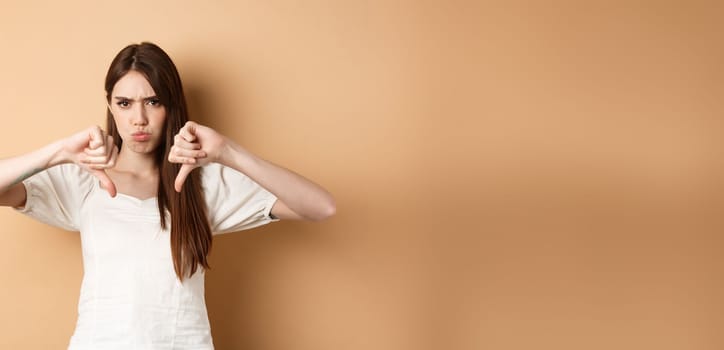 Absolutely no. Disappointed young woman frowning and showing thumbs down, express dislike and negative emotions, bad feedback, standing on beige background.
