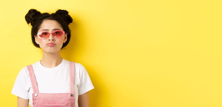 Summer and fashion concept. Bored asian teen girl in sunglasses pouting, standing moody on yellow background.