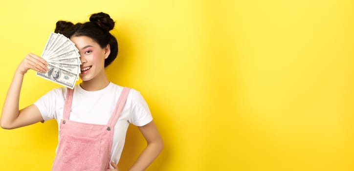 Shopping. Beautiful asian female model with glamour makeup, cover half of face with money dollar bills, smiling sensual at camera, yellow background.