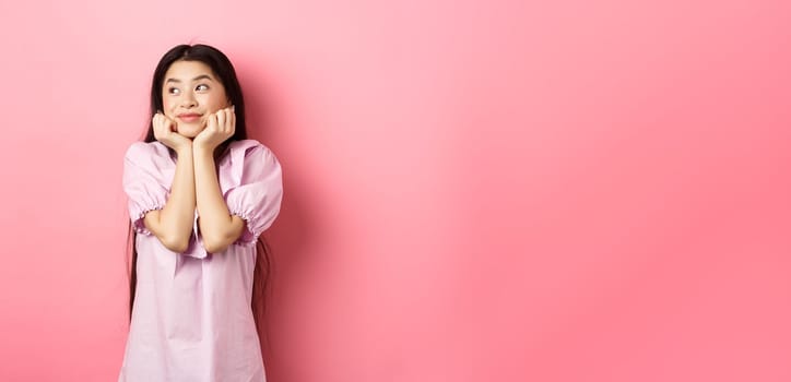 Dreamy romantic girl looking aside at logo and smiling, thinking of something beautiful, standing on pink background.