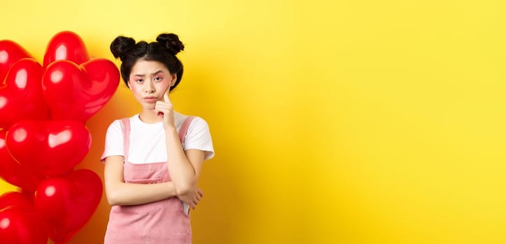 Valentines day concept. Unamused and bored asian girlfriend staring skeptical at camera, waiting for something near red hearts balloons, yellow background.