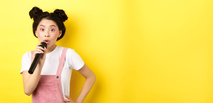 Cute asian teen girl with bright makeup, singing in microphone karaoke, standing against yellow background.