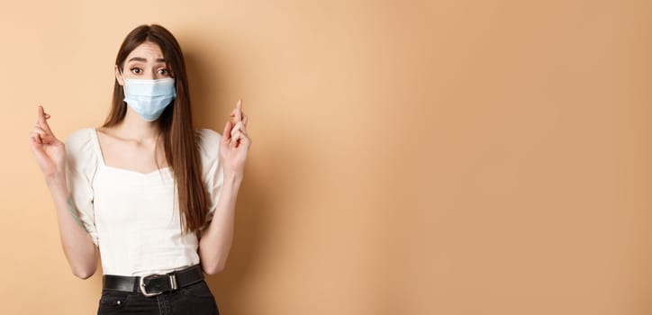 Covid-19 and lifestyle concept. Hopeful girl in face mask praying with fingers crossed, looking with hope at camera, standing on beige background.