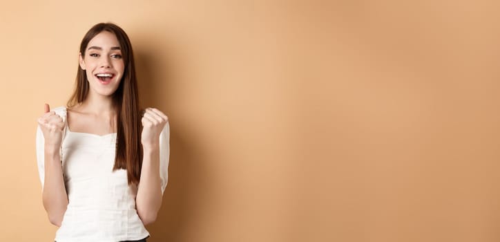 Happy smiling girl celebrating victory, making fist pump and say yes, achieve goal, triumphing or getting motivated, standing on beige background.