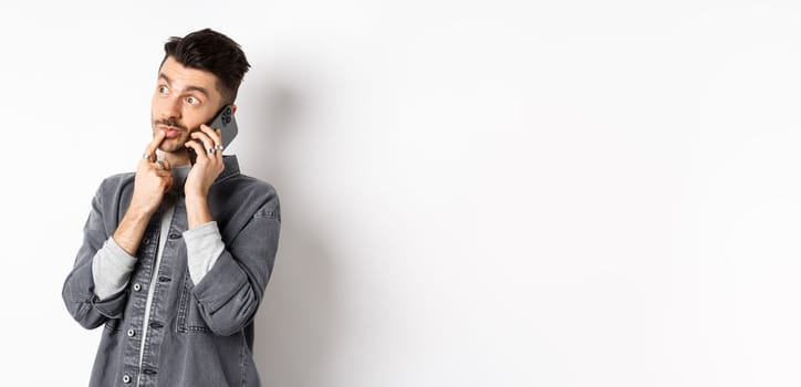 Image of pensive handsome man talking on phone and thinking, making choice while making takeout order, standing against white background.